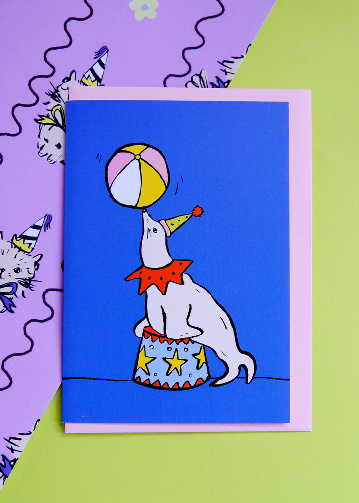 Card with a dog in a clown outfit playing with a ball on a blue background, on a purple and green checkered surface.
